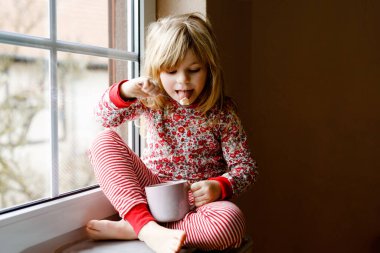 Küçük anaokulu kızı elinde şekerlemeli sıcak çikolata bardağıyla. Pijamalı mutlu çocuk kışın pencerenin yanında tatlı kakao içiyor. Noel 'in samimi aile kutlaması.