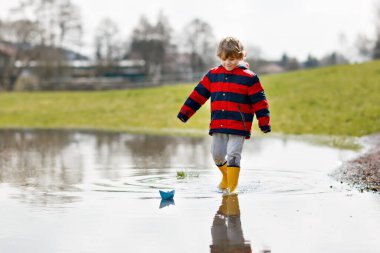 Sarı çizmeli mutlu çocuk baharda ya da sonbahar günü büyük su birikintisinin yanında kağıt gemi teknesiyle oynuyor.