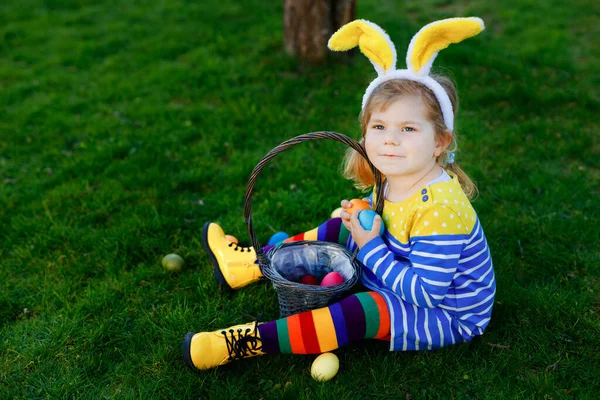 Jolie petite fille tout-petit assis sur l'herbe verte et porter des oreilles de lapin, s'amuser avec des œufs traditionnels de couleur de Pâques chassent par une chaude journée ensoleillée, en plein air. Joyeux enfant célébrant des vacances en famille. — Photo