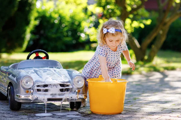 Linda niña hermosa lavando un gran coche de juguete viejo en el jardín de verano, al aire libre. Feliz niño sano coche de limpieza con agua y jabón, divertirse con salpicaduras y jugar con esponja. — Foto de Stock