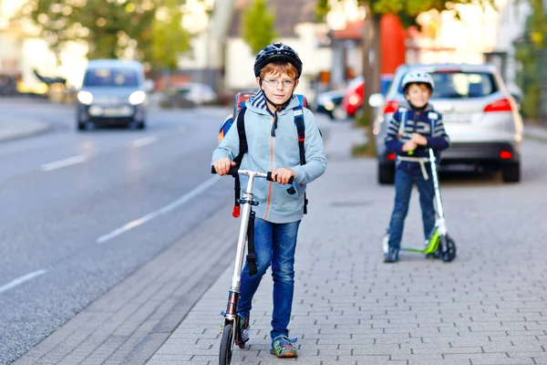 Güneşli bir günde, ellerinde scooterla şehirde gezen kasklı iki okul çocuğu. Renkli giysiler içinde okula bisikletle giden mutlu çocuklar.. — Stok fotoğraf