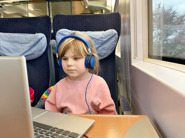 Милая маленькая дошкольница сидит в поезде и смотрит телевизор на ноутбуке во время переезда. Счастливый ребенок едет в семейный отпуск по железной дороге. Девушка с ноутбуком и наушниками — стоковое фото