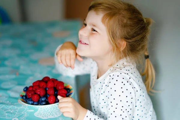 Aranyos kis óvodás lány, aki friss málnát és áfonyát eszik. Boldog gyermek málnát és áfonyát kóstol. Egészséges étel, gyermekkor és fejlődés. Boldog gyerek otthon vagy gyerekszobában. — Stock Fotó
