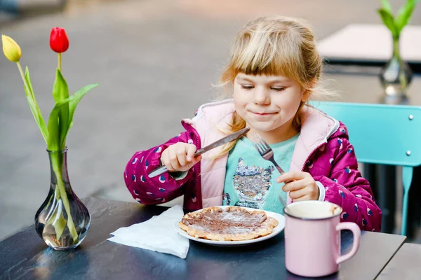 屋外カフェレストランに座ってかわいい就学前の女の子。幸せな健康な子供は甘いワッフルかクレープを食べ、ナイフを使用し、晴れた日にフォークを学ぶ。ホットチョコレートを飲む子供 — ストック写真