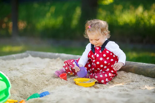 Симпатичная малышка играет на песке на открытой площадке. Ребенок в красных брюках веселится в солнечный теплый летний день. Ребенок с разноцветными песчаными игрушками. Здоровый активный ребенок на открытом воздухе играет в игры — стоковое фото