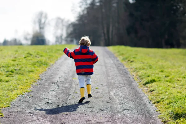 Little kid boy wearing yellow rain boots and walking on nature path on warm sunny spring day. — Stock Photo, Image