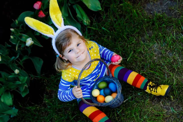 Söt liten småbarn flicka med kaninöron har kul med traditionella påskägg jakt på varm solig dag, utomhus. Glad barn firar familj kristna semester med korg med färgade ägg — Stockfoto