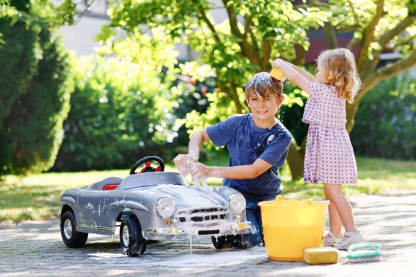 Duas crianças felizes lavando grande carro de brinquedo velho no jardim de verão, ao ar livre. Irmão menino e irmã mais nova menina de limpeza carro com sabão e água, se divertindo com espirrar e brincar com esponja. — Fotografia de Stock