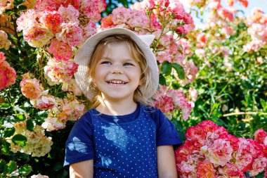 Çiçek açan gül bahçesinde küçük bir kızın portresi. Yaz güneşli bir günde parkta güllerle ve çiçeklerle eğlenen sevimli güzel bir çocuk. Mutlu gülümseyen bebek.