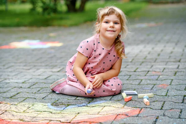 Little preschool girl painting rainbow with colorful chalks on ground on backyard. Positive happy toddler child drawing and creating pictures. Creative outdoors activity in summer. — Stock Photo, Image