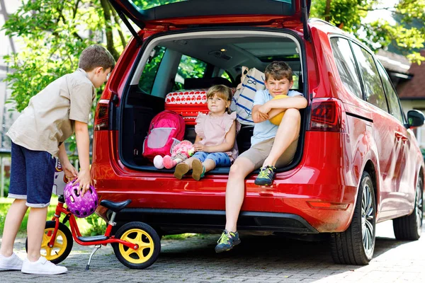 Drei Kinder, zwei Jungen und ein Vorschulmädchen sitzen im Kofferraum des Autos, bevor sie mit ihren Eltern in die Sommerferien fahren. Glückliche Kinder, Geschwister, Geschwister mit Koffern und Spielzeug auf Reisen — Stockfoto