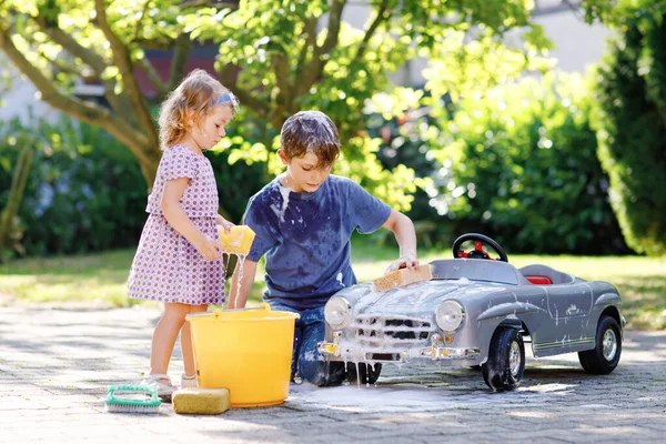 Zwei glückliche Kinder waschen im Sommergarten im Freien ein großes altes Spielzeugauto. Bruder Junge und kleine Schwester Kleinkind Mädchen reinigen Auto mit Seife und Wasser, haben Spaß beim Planschen und Spielen mit Schwamm. — Stockfoto