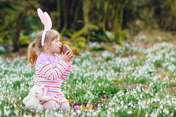 Liten flicka med påskharen öron gör ägg jakt i vårskogen på solig dag, utomhus. Söt glad barn med massor av snödroppar blommor, stor choklad ägg och färgade ägg. — Stockfoto