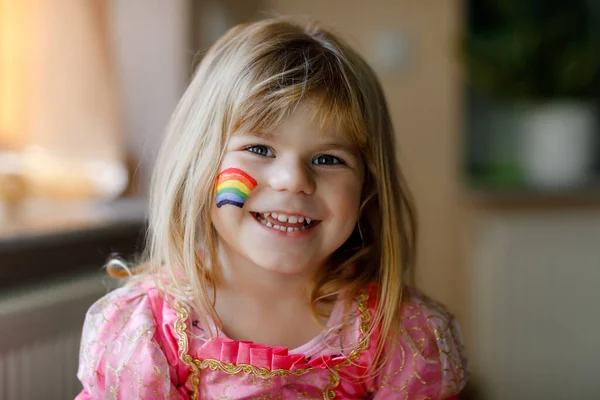 Retrato de linda niña pequeña con arco iris pintado con colores coloridos en la cara. Niño feliz con signo de diversidad y paz en el mundo. — Foto de Stock