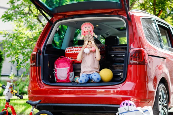 Menina pré-escolar bonito sentado no porta-malas do carro antes de sair para férias de verão com os pais. Criança ativa feliz com malas, bicicleta e brinquedos em viagem familiar, viagem de carro — Fotografia de Stock