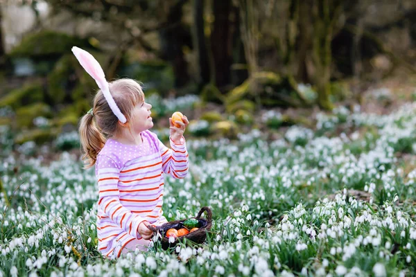 一个小女孩，带着复活节兔子的耳朵，在阳光明媚的春天的森林里打猎。可爱快乐的孩子,有许多雪花和彩蛋.春天，基督教节日的概念. — 图库照片