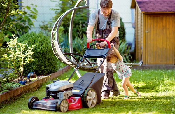 Happy little toddler girl and middle-aged father with lawn mower. Family, daughter, preschool child and dad cut the lawn. Portrait of family working in garden, trimming grass. Garden works in summer. — Stock Photo, Image