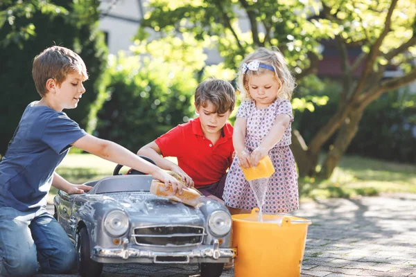 Três crianças felizes lavando grande carro de brinquedo velho no jardim de verão, ao ar livre. Dois meninos e pequena menina limpando carro com água e sabão, se divertindo com espirrar e brincar com esponja. — Fotografia de Stock