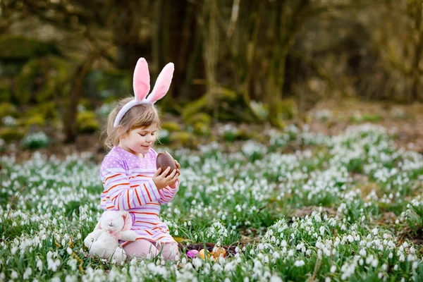 Liten flicka med påskharen öron gör ägg jakt i vårskogen på solig dag, utomhus. Söt glad barn med massor av snödroppar blommor, stor choklad ägg och färgade ägg. — Stockfoto