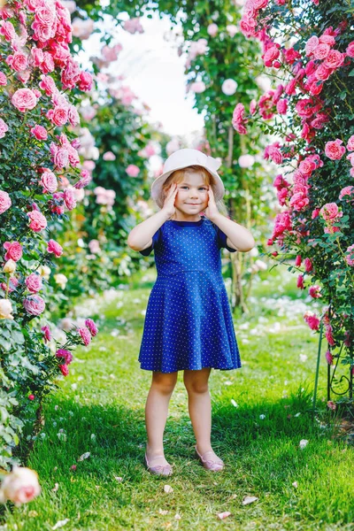 Portrét malé batolátko v rozkvetlé růžové zahradě. Roztomilé krásné krásné dítě baví s růžemi a květinami v parku v létě slunečný den. Šťastné usměvavé dítě. — Stock fotografie
