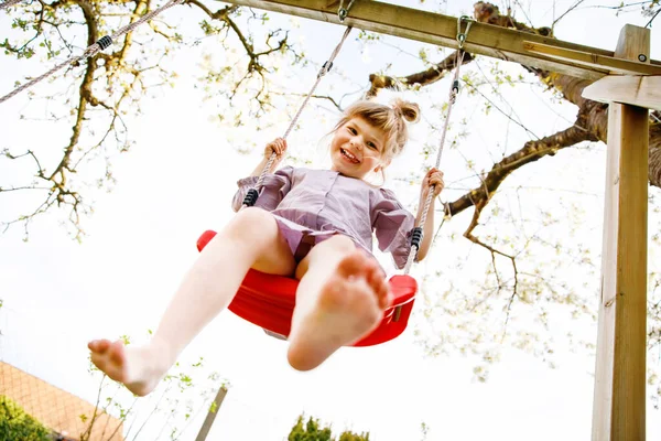 Glad liten småbarn flicka som har kul på swing i hushållsträdgården. Leende positiva friska barn svänger på solig dag. Förskoleflickan skrattar och gråter. Aktiv fritid och aktivitet utomhus. — Stockfoto