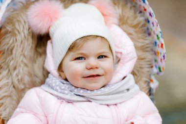 Tatlı küçük güzel kız bebek arabasında oturuyor ya da soğuk sonbaharda, kış ya da bahar günü bebek arabasında. Sıcak giysili, şık bir bebek ceketi ve şapkası olan mutlu bir çocuk. Kar yağıyor.