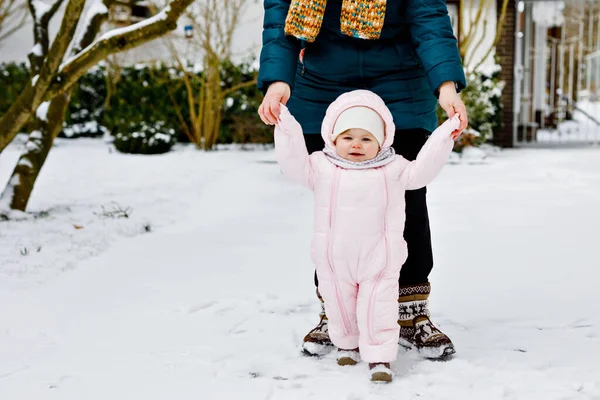 Очаровательная маленькая девочка делает первые шаги на улице зимой через снег. Милый малыш учится ходить. Мать держит ребенка под рукой. Дочь и мама ходят вместе. Счастливая семья — стоковое фото