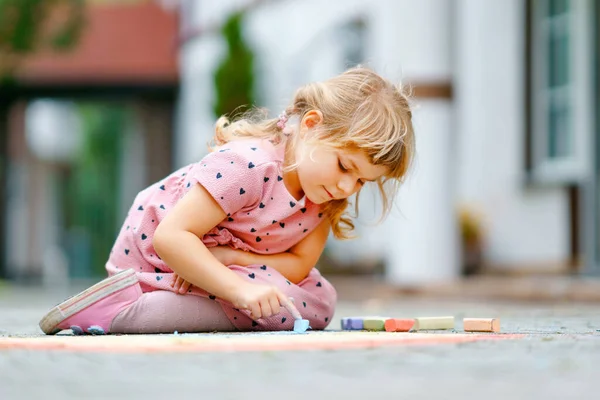 Μικρό κορίτσι προσχολικής ζωγραφικής ουράνιο τόξο με πολύχρωμα κιμωλίες στο έδαφος στην πίσω αυλή. Θετικό, χαρούμενο παιδί ζωγραφίζει και δημιουργεί εικόνες. Δημιουργική υπαίθρια δραστηριότητα το καλοκαίρι. — Φωτογραφία Αρχείου