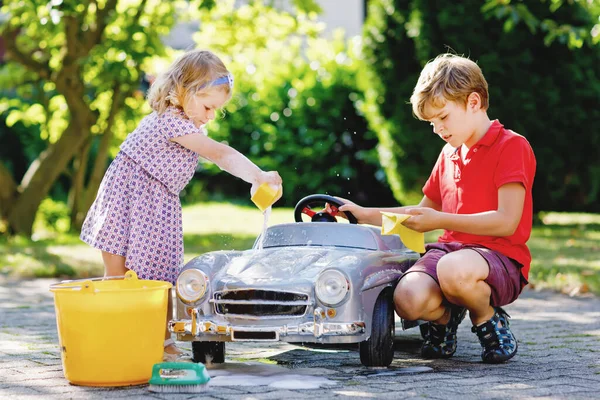 Deux enfants heureux laver grande vieille voiture jouet dans le jardin d'été, à l'extérieur. Frère garçon et petite sœur tout-petit fille nettoyage voiture avec du savon et de l'eau, s'amuser avec éclaboussures et jouer avec éponge. — Photo