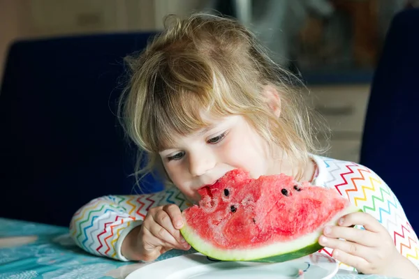 Mała przedszkolanka jedząca świeży czerwony arbuz. Maluch gryzie dojrzałe zdrowe owoce. Kid korzystających przekąski w kuchni domowej, w pomieszczeniach. — Zdjęcie stockowe