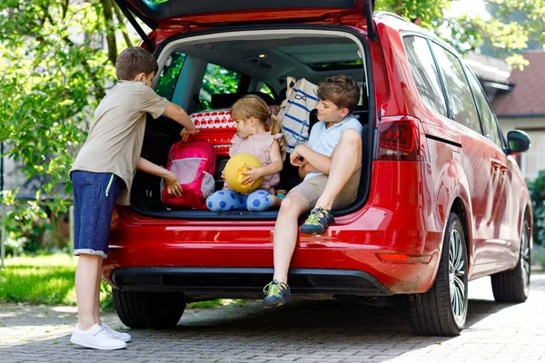 Tres niños, dos niños y una niña preescolar sentados en el maletero del coche antes de salir de vacaciones de verano con los padres. Niños felices, hermanos, hermanos y hermanas con maletas y juguetes de viaje —  Fotos de Stock