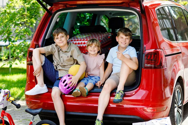 Trois enfants, deux garçons et une fille d'âge préscolaire assis dans le coffre de la voiture avant de partir pour des vacances d'été avec les parents. Enfants heureux, frères et sœurs, frères et sœurs avec valises et jouets en voyage — Photo