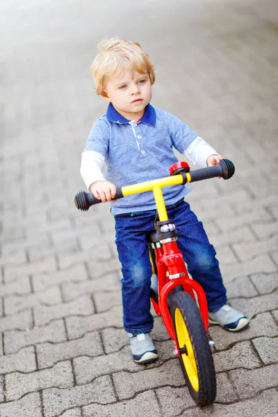 Active blond kid boy in colorful clothes driving balance and learners bike or bicycle in domestic garden. Toddler child dreaming and having fun on warm summer day. outdoors movement game for children — Stock Photo, Image