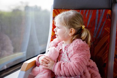 Trende oturan ve hareket halindeyken pencereden dışarı bakan tatlı küçük kız. Sevimli, sağlıklı, peluş oyuncağı elinde tutan bebek. Gülümseyen çocuk aile tatiline gidiyor demiryolu ile