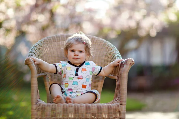 Carina bambina seduta su una grande sedia in giardino. Bella felice bambino sorridente con albero di magnolia rosa in fiore sullo sfondo. Bambino sano godendo della stagione primaverile. — Foto Stock