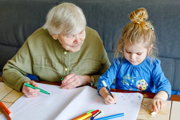 Hermosa niña y abuela dibujando juntos cuadros con plumas de fieltro en casa. Lindo niño y mujer mayor divirtiéndose juntos. Familia feliz en el interior — Foto de Stock