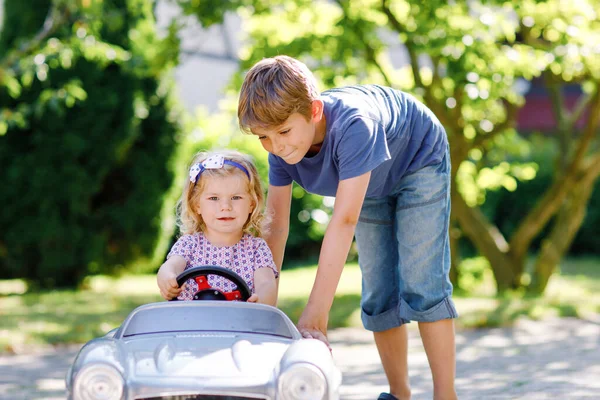 Dos niños felices jugando con un gran coche de juguete viejo en el jardín de verano, al aire libre. Chico conduciendo coche con la niña, hermana linda dentro. Riendo y sonriendo niños. Familia, infancia, concepto de estilo de vida. — Foto de Stock