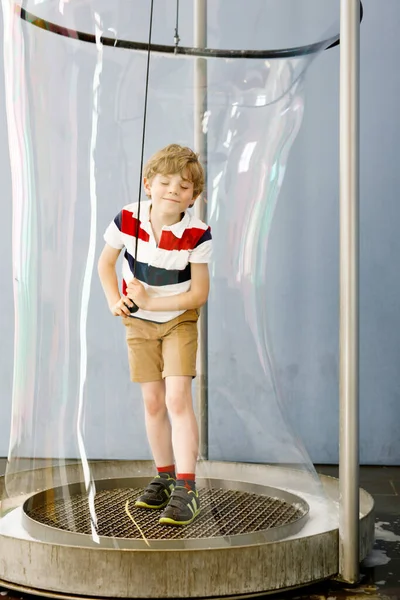 Маленький блондин, играющий с огромными мыльными пузырями в помещении. Счастливый здоровый улыбающийся ребенок веселится с экспериментами — стоковое фото