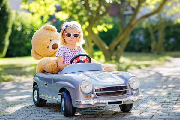 Pequeña niña adorable conduciendo un gran coche de juguete vintage y divirtiéndose jugando con un oso de peluche, al aire libre. Precioso niño feliz y saludable disfrutando de un cálido día de verano. Sonriente niño impresionante en gaden — Foto de Stock