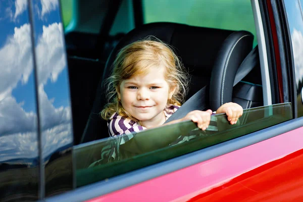 Förtjusande liten flicka sitter i bilbarnstolen och tittar ut genom fönstret på natur och trafik. En liten unge som reser med bil. Barnsäkerhet på vägen. Familjeresa och semester på sommaren — Stockfoto