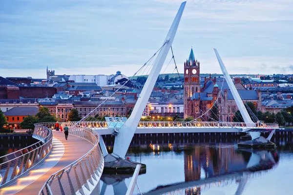 Derry, Ireland. Illuminated Peace bridge in Derry Londonderry, City of Culture, in Northern Ireland with city center at the background. Night cloudy sky with reflection in the river at the dusk — Stock Photo, Image