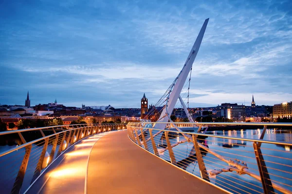 Дерри, Ирландия. Освещённый мост Мира в Дерри-Лондондерри, городе культуры, в Северной Ирландии с центром города на заднем плане. Ночное облачное небо с отражением в реке в сумерках — стоковое фото