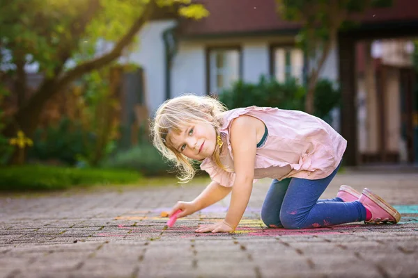 Little preschool girl painting with colorful chalks flowers on ground on backyard. Positive happy toddler child drawing and creating pictures on asphalt. Creative outdoors children activity in summer. — Stock Photo, Image