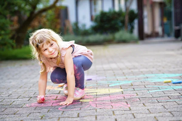 Kleine kleuter meisje schilderen met kleurrijke krijtjes bloemen op de grond op de achtertuin. Positief gelukkig peuter kind tekenen en het creëren van foto 's op asfalt. Creatief buitenshuis kinderactiviteiten in de zomer. — Stockfoto