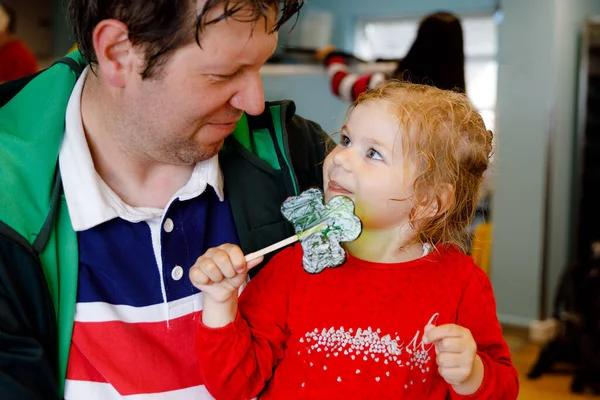 Cute toddler girl and father with cloverleaf lollipop in Irish pub or cafe. Child and dad, young man resting with parents and eating unhealthy sweets. Family making vacation in Ireland. — Stock Photo, Image