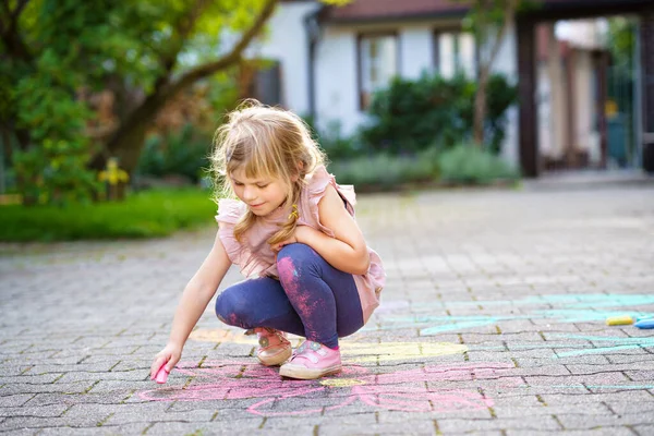 Little preschool girl painting with colorful chalks flowers on ground on backyard. Positive happy toddler child drawing and creating pictures on asphalt. Creative outdoors children activity in summer. — Stock Photo, Image