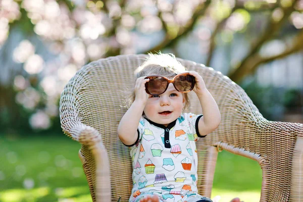 Мила маленька дівчинка сидить на великому стільці в саду. Прекрасний щасливий усміхнений малюк з розквітлим рожевим магнолієвим деревом на фоні. Здорова дитина насолоджується весняним сезоном . — стокове фото