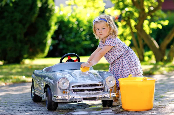 Mignon magnifique tout-petit fille lavage grande vieille voiture jouet dans le jardin d'été, en plein air. Heureux petit enfant sain nettoyage de voiture avec du savon et de l'eau, s'amuser avec éclaboussures et jouer avec l'éponge. — Photo