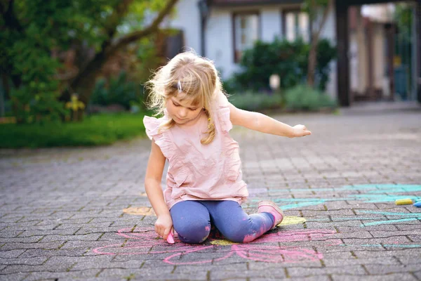 Kleine kleuter meisje schilderen met kleurrijke krijtjes bloemen op de grond op de achtertuin. Positief gelukkig peuter kind tekenen en het creëren van foto 's op asfalt. Creatief buitenshuis kinderactiviteiten in de zomer. — Stockfoto