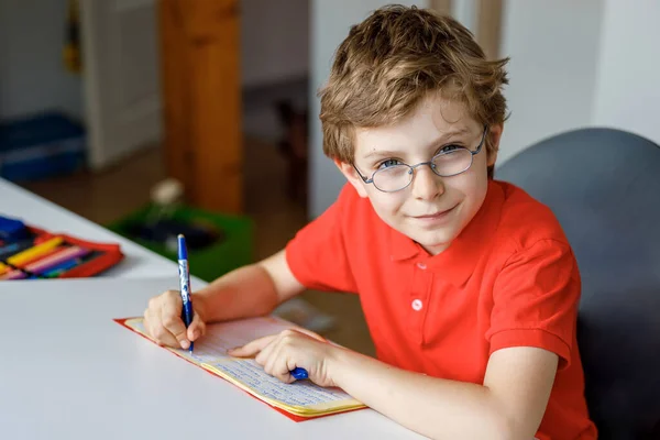 Portret van een kleine jongen met een bril thuis die huiswerk maakt, schrijft en leert. Klein kind aan het sporten, binnen. Basisschool en -onderwijs, thuisonderwijs. — Stockfoto